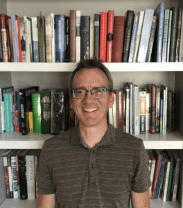 Charles Perrin, a man wearing a green collared shirt, stands in front of a bookshelf smiling at the camera