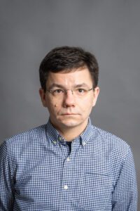 Janis Juzefovics, a man with short brown hair and glasses, wearing a blue and white checked shirt, looks at the camera with a neutral expression.