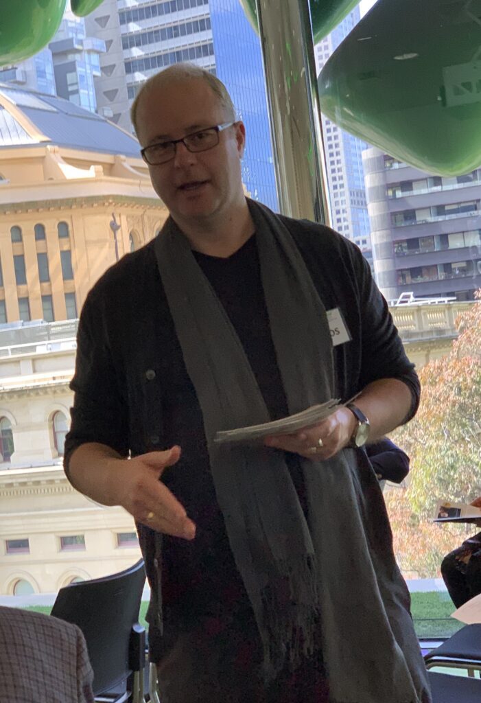 Dr. Delaney Skerret, a man with blond hair and glasses, wearing a black shirt and a long green scarf, speaks while gesturing downward with his right hand