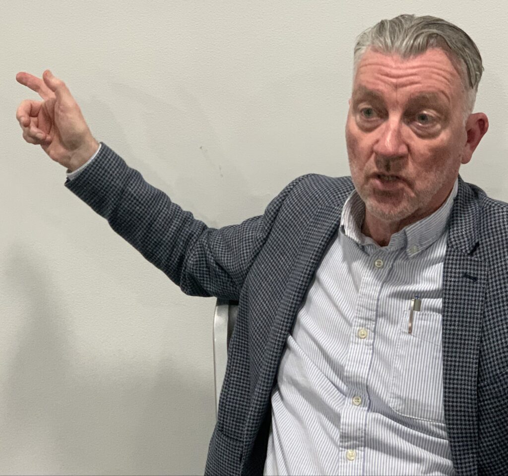 Ron Popenhagen, a man with short gray hair and weearinng a dark gray jacket over a blue shirt, gestures emphatically with his right arm as he speaks, with the camera cropped in close