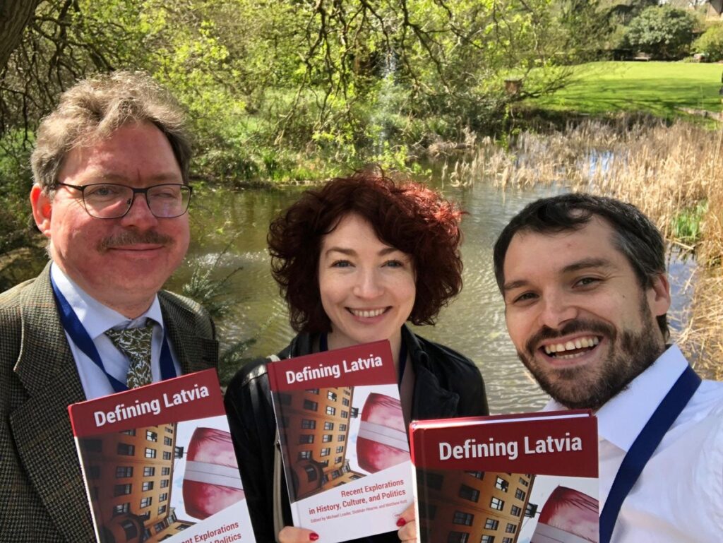 Two men and a woman standing in front of a pond. Each hold a copy of a book entitled "Defining Latvia"