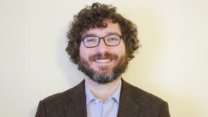 A man with curly hair, glasses, and a beard smiles at the camera.